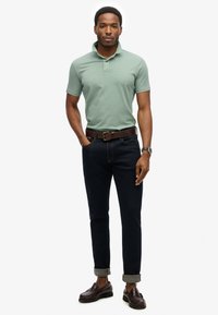 Polo verde maniche corte, jeans di denim scuro arrotolati alle caviglie, cintura in pelle marrone scuro e scarpe scure con finitura texture.