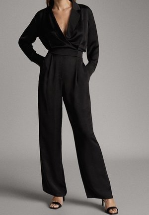Woman wearing a black long-sleeve wrap blouse and high-waisted wide-leg pants paired with black open-toe heels, standing with hands in pockets.