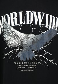 Schwarzes T-Shirt mit einem großen, grauen Adler-Graphic, detaillierten Federn, Blitzmotiven und dem Text "WORLDWIDE TOUR" darüber.
