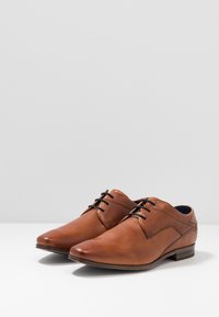 Chaussures habillées en cuir marron avec une texture lisse, bout pointu, lacets noirs et un petit talon empilé. Présente un détail de couture latérale subtil.