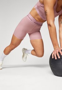 Pessoa em roupa ativa rosa da New Balance faz prancha com as mãos sobre uma bola medicinal preta, usando ténis e meias brancas.