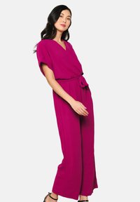 Magenta jumpsuit met een V-hals, korte mouwen en een strikceintuur. Vloeiende wijde pijpen, zachte stof, met zijsplitten voor extra beweging.