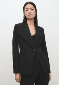 Czarna marynarka na jeden guzik, dopasowanego kroju, z wyciętymi klapami i przednimi kieszeniami. Stylizowana z czarnym topem i eleganckim designem.