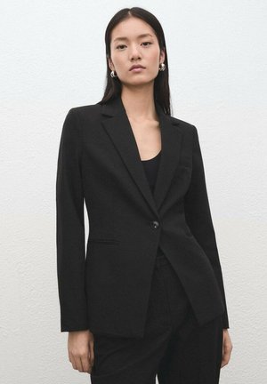 Czarna marynarka na jeden guzik, dopasowanego kroju, z wyciętymi klapami i przednimi kieszeniami. Stylizowana z czarnym topem i eleganckim designem.