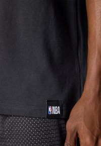 Črna bombažna majica s kratkimi rokavi s kupljenim NBA značko na robu; ima gladko teksturo in preprost, klasičen dizajn.