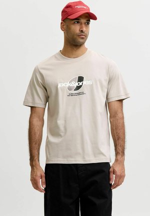 Mand iført en beige Jack & Jones T-shirt med sort tekst og en rød kasket, stående foran en ensfarvet lys baggrund.