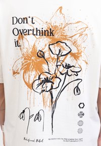 Wit katoenen T-shirt met oranje en zwarte bloemprint en de tekst "Don't Overthink it." Bevat merknaam en kleine grafische details.