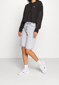 Felpa corta nera con logo, maniche lunghe con strass. Pantaloncini di jeans azzurri, sneakers bianche con dettagli grigi e calzini alla caviglia.