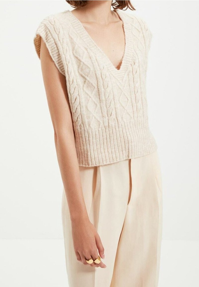 Gilet en tricot beige avec un col en V, des manches courtes et un ourlet côtelé. Porté par-dessus un pantalon beige, mettant en valeur un motif de tricot texturé.