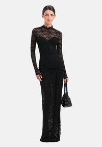 Zwarte kanten jurk met lange mouwen, een aansluitend silhouet, een gestructureerd bloemmotief, een sweetheart halslijn en zijwaartse drapering. Model houdt een kleine zwarte tas vast.