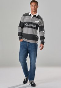 Rugby-style shirt in gray and black stripes with white collar, featuring a Guinness logo. Worn with blue jeans and black sneakers.