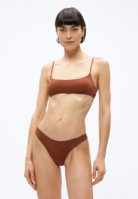 Sieviete ar īsiem tumšiem matiem un riņķveida auskariem, tērpusies brūnā bikini ar plānām siksnām, stāv pie vienkārša balta fona.