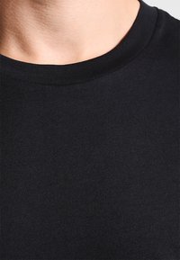 Detailní záběr na krk a horní část hrudníku osoby v jednoduchém černém tričku s kulatým výstřihem, na bílém pozadí.