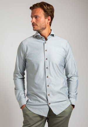 Lichtblauwe blouse met lange mouwen, knoopsluiting en een subtiel gestreept patroon, gecombineerd met olijfgroene broek.