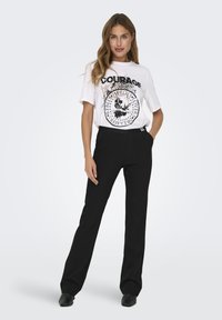 Witte t-shirt met zwart grafisch print en tekst "COURAGE." Zwarte, op maat gemaakte broek met een strakke pasvorm en voortassen.
