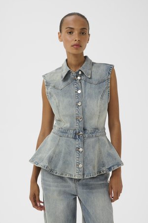 Frau trägt ärmelloses, geknöpftes, hellblaues Denim-Peplum-Oberteil mit Kragen, kombiniert mit passenden hellblauen Denim-Jeans, vor weißem Hintergrund.