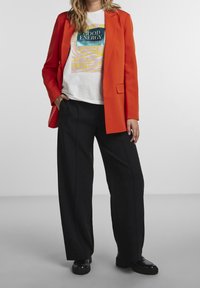 Blazer orange sur un t-shirt blanc à motif avec le message « GOOD ENERGY » et un pantalon noir à jambes larges ; bottes noires brillantes.