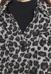 Leopardikuvioinen takki harmaana ja mustana, pehmeästä kankaasta valmistettu. Siinä on kauluksellinen muotoilu, mustat napit ja rintataskut.