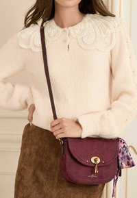 Borsa a tracolla in suede bordeaux con hardware dorato e un nastro di seta decorato. Texture morbida, forma curvata e dettagli di cucitura visibili.