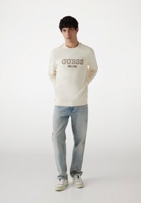 Felpa crema con logo "GUESS" in marrone, abbinata a jeans azzurri chiari e sneaker bianche, caratterizzata da una vestibilità rilassata e polsini a coste.