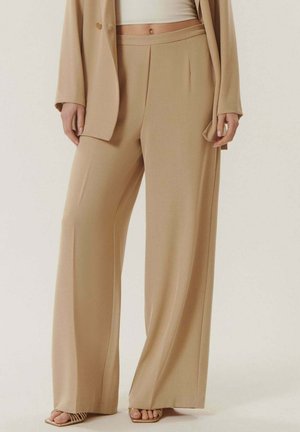 Femme portant un pantalon beige à jambes larges assorti d'un blazer, un crop top blanc et des sandales à talons ouvertes, debout devant un fond uni.