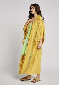 Geel gefranjert kimono-stijl buitengewear gelaagd over een groene jurk, met platte bruine sandalen aan de voeten, en een losse pasvorm.
