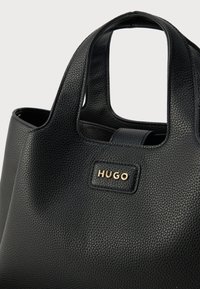 Geantă tote din piele neagră cu textură granulată, cu mânere scurte și o etichetă a brandului aurie inscripționată „HUGO” pe lateral.