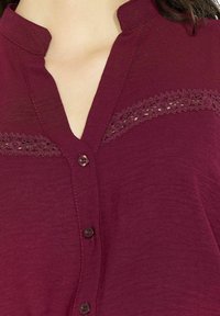 Camicia bordeaux con bottoni, caratterizzata da un colletto alla mandarino, dettagli in pizzo lungo il petto e tessuto testurizzato con vestibilità rilassata.
