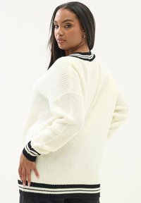 Biały dzianinowy sweter z czarnym i białym prążkowanym kołnierzem i mankietami. Luźny krój z teksturowanej tkaniny i długimi rękawami.