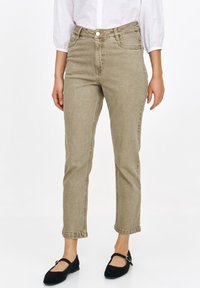 Beżowe jeansy z wysokim stanem o dopasowanym kroju i subtelnej teksturze, z przednimi kieszeniami i klasycznym zapięciem na guzik. Nosi je w połączeniu z czarnymi balerinami.
