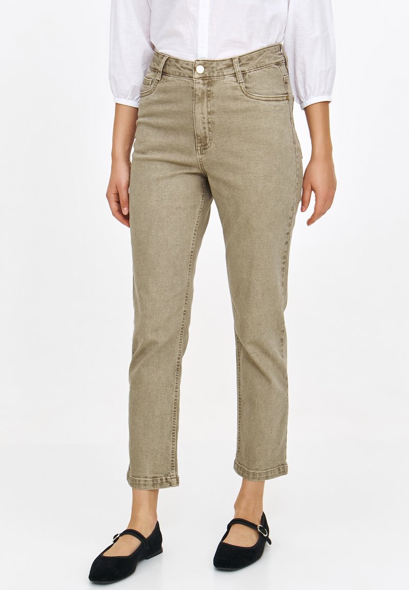 Beżowe jeansy z wysokim stanem o dopasowanym kroju i subtelnej teksturze, z przednimi kieszeniami i klasycznym zapięciem na guzik. Nosi je w połączeniu z czarnymi balerinami.