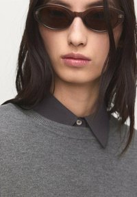 Gray sweater with a round neckline, layered over a dark gray collared shirt. Model wears brown oval sunglasses. Smooth textures, minimal design.