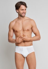 Smilende mand uden trøje iført hvide underbukser, stående mod en ensfarvet lys grå baggrund, og ser mod sin venstre side.