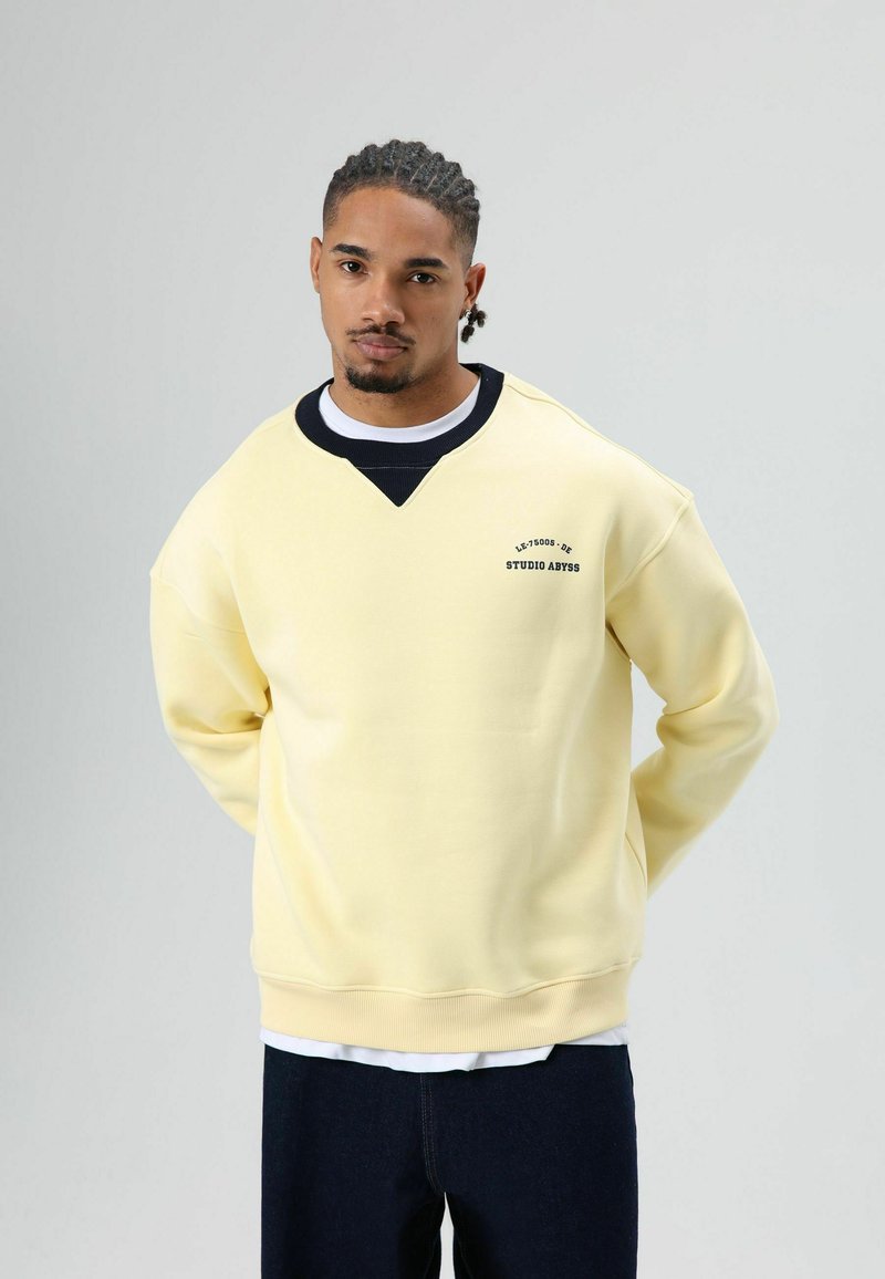 Lichtgele sweatshirt met een zwarte halslijn en een logo op de borst. Heeft een losse pasvorm en geribbelde manchetten. Draagt over een wit shirt.