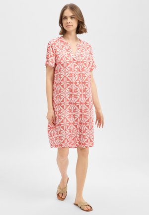 Jonge vrouw draagt een koraalroze en witte jurk met patroon tot over de knie en beige platte sandalen, staand voor een effen lichte achtergrond.