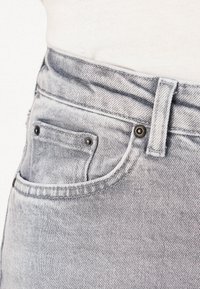 Gros plan sur un jean en denim gris clair montrant une poche avant, une petite poche, une passants de ceinture et des rivets métalliques sur fond blanc.