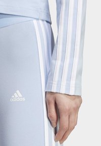 Primer plano de una mano descansando cerca de unos pantalones y una chaqueta de entrenamiento Adidas de color azul claro con detalles de tres franjas blancas.