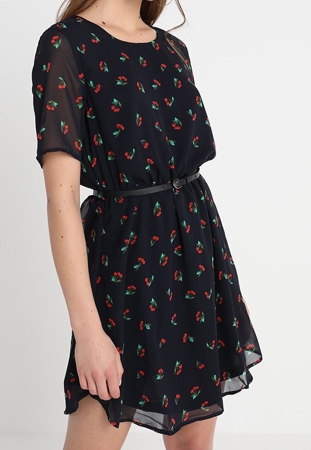 Robe noire à manches courtes avec motif cerises rouges, cintrée à la taille par une fine ceinture noire, portée par une personne debout.