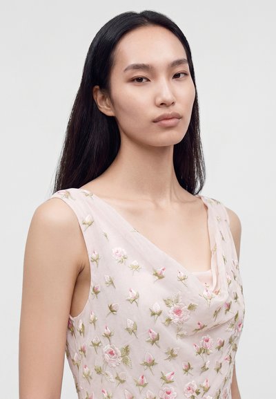 Femme aux longs cheveux noirs portant une robe sans manches rose pâle avec des motifs floraux brodés roses et verts, regardant calmement devant elle.