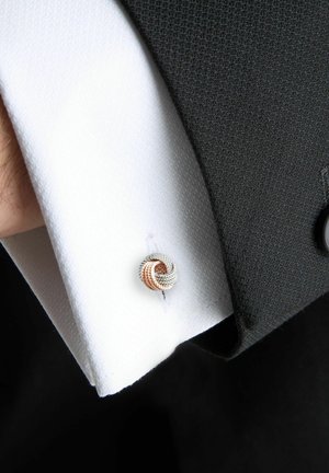 Weißer, strukturierter Hemdmanschetten mit einer silbernen und roségold gedrehten Knotenmanschettenknopf, teilweise bedeckt von einem schwarzen Anzugsärmel.