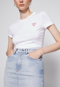 Balta prigludusi marškinėliai su raudona trikampio „GUESS“ logotipu krūtinėje, derinami su šviesiai mėlynos spalvos džinsiniu sijonu su priekinėmis kišenėmis.