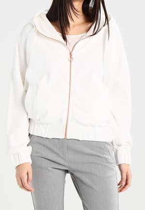 Kobieta ubrana w biały rozpinany na zamek kaptur z kapturem i prążkowanymi mankietami, w połączeniu z jasnoszarymi spodniami, stojąca na białym tle.