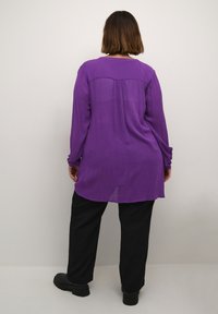 Blouse violette à manches longues en tissu léger, avec un pli au dos et un ourlet arrondi. Portée avec un pantalon noir et des bottes.