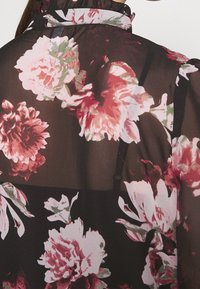 Blouse noire transparente avec de grands motifs floraux vibrants en rose et blanc. Dotée d'un col à volants et d'une conception à bretelles spaghetti.