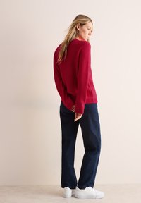 Rode gebreide trui met een losse pasvorm, gecombineerd met marineblauwe broek en witte sneakers. De trui heeft een gestructureerd patroon en ribgebreide manchetten.