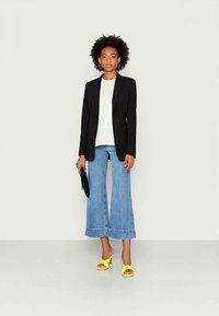 Blazer preto, blusa branca, calças jeans azuis de perna larga e sandálias de salto amarelas. Modelo segura uma clutch preta. Cores sólidas, corte ajustado.