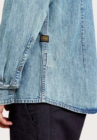 Jasnoniebieska kurtka dżinsowa z zapięciem na guziki na mankiecie, bocznym rozcięciem oraz czarną metką z napisem "G-Star Originals" na dole.