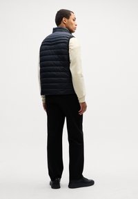 Μαύρο γιλέκο τύπου πούφτερ με ψηλό λαιμό, υφή και εφαρμοστή σχεδίαση. Φορεμένο πάνω από ένα ανοιχτόχρωμο μακρυμάνικο μπλουζάκι και μαύρο παντελόνι.