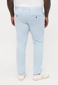 Hellblaue Hose mit glatter Textur, taillierter Form und hinteren Taschen mit schwarzen Knöpfen. Kombiniert mit weißen Sneakers.