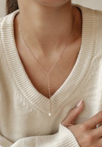 Różowo-złoty naszyjnik z długim łańcuchem i małym akcentem perłowym, noszony na kremowym swetrze dzianinowym z głębokim dekoltem w kształcie litery V.